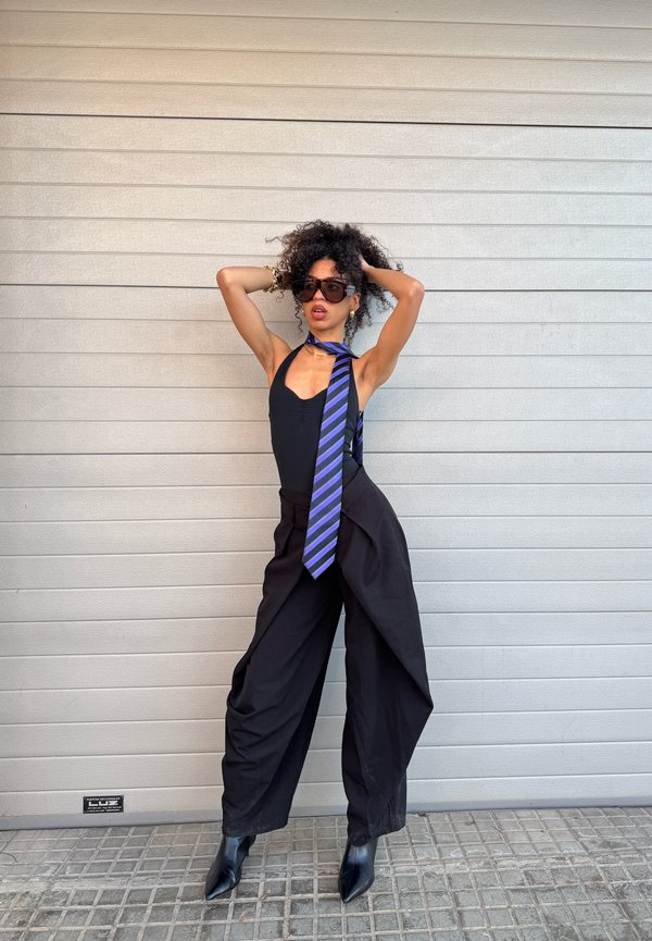 Kvinde med krøllet hår iført solbriller, en sort ærmeløs top, sorte bukser med vide ben, sorte støvler og et blåt striped slips, der poserer mod en grå væg.
