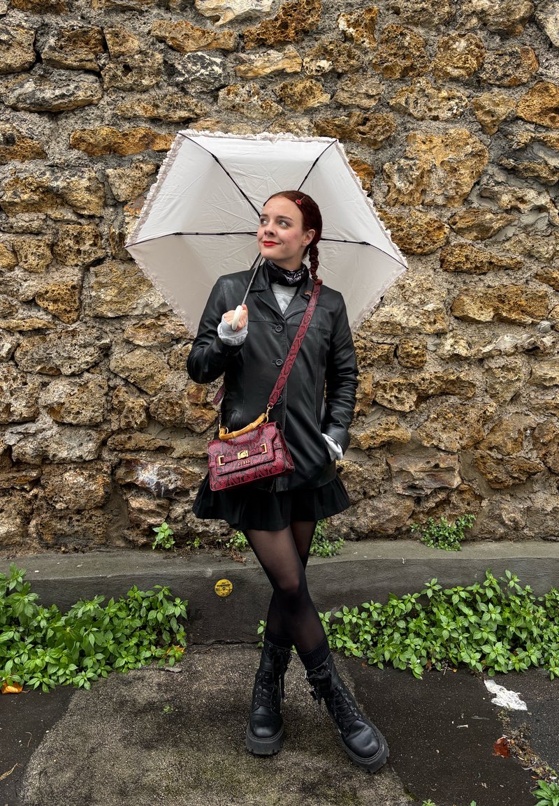 Manteau en cuir noir, parapluie blanc, sac à main rouge à motifs, jupe courte noire, collants noirs, bottes noires épaisses, devant un mur en pierre.