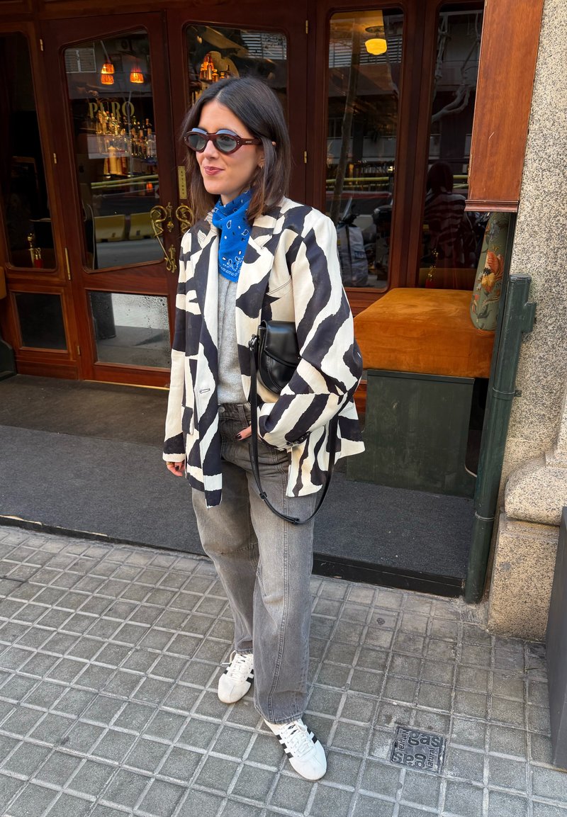 Femme portant une veste zébrée, un jean gris, des baskets blanches, un bandana bleu et des lunettes de soleil, debout sur un trottoir en ville devant une porte en bois.