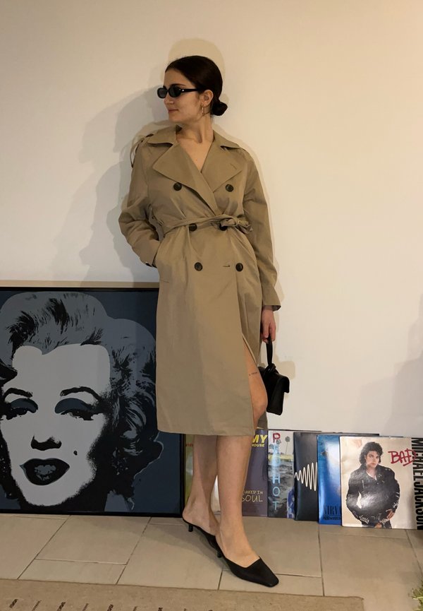 Žena v béžovom trenčkoté, čiernych slnečných okuliaroch a na podpätkoch stojí vedľa steny s umeleckým dielom Marilyn Monroe a vinylovými gramofónovými platňami.