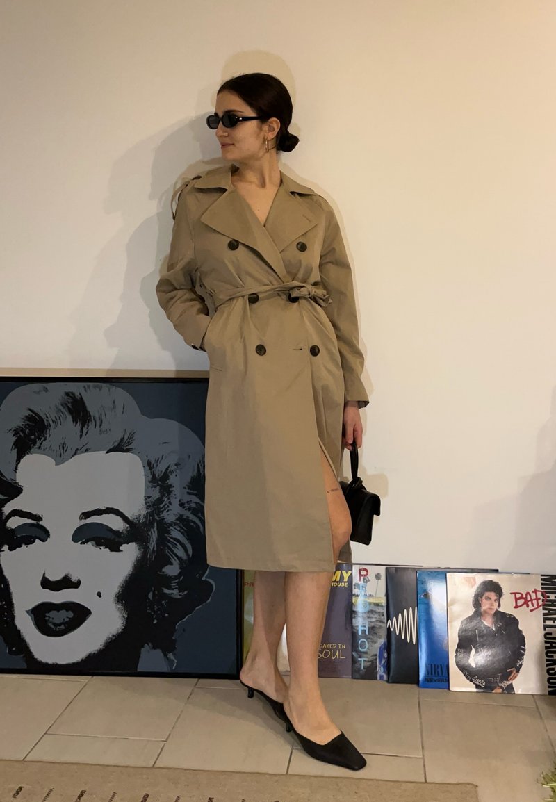 Femme portant un trench-coat beige, des lunettes de soleil noires et des talons, debout à côté d'un mur avec une œuvre d'art de Marilyn Monroe et des disques vinyles.