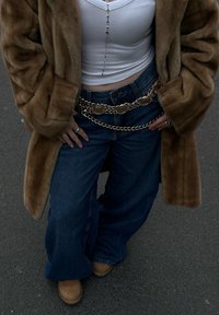 Καφετί συνθετικό παλτό από γούνα, λευκό κοντό τοπ, φαρδιοί τζιν σε μπλε απόχρωση, χρυσός αλυσίδας ζώνη, καφέ μπότες. Τα αξεσουάρ περιλαμβάνουν δαχτυλίδια και μια κολιέ με χάντρες.