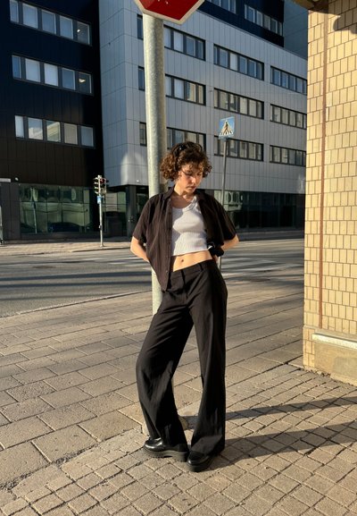 Pantalones anchos y casuales de color negro con un ajuste holgado, combinados con una camiseta corta de rayas blancas y un camisa negra de botones corta, llevados con zapatos negros gruesos.