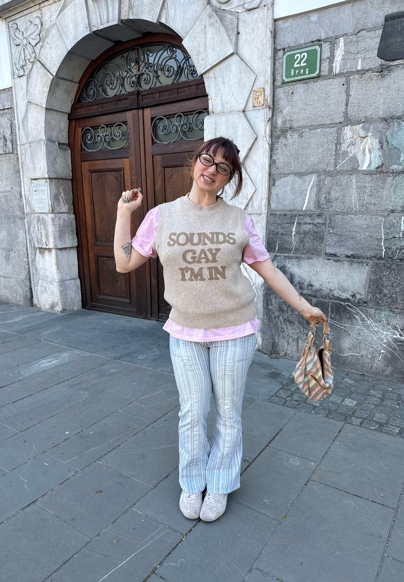 Personne souriante avec des lunettes, portant un gilet en maille « SOUNDS GAY I'M IN », chemise rose, pantalon rayé, tenant un sac rayé près de portes en bois cintrées.