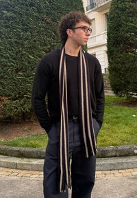 Άνδρας με σγουρά μαλλιά και γυαλιά, φορώντας μαύρο πουλόβερ, ριγέ κασκόλ και σκούρο παντελόνι, στέκεται έξω με τα χέρια στις τσέπες.