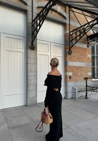 Zwarte geribbelde off-shoulder jurk met lange mouwen en een uitsnijding, gecombineerd met een geweven handtas. Achtergrond van een bakstenen en grijs gebouw.