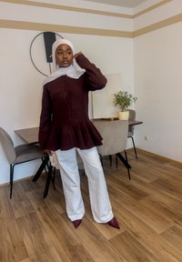 Haut péplum bordeaux boutonné, pantalon blanc à jambes larges, et chaussures rouges pointues. Hijab blanc, accessoires minimalistes, cadre intérieur avec une plante.
