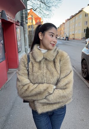 Joven con el cabello largo y oscuro, vestida con una chaqueta de piel color canela, de pie en una acera de la ciudad con los brazos cruzados, mirando hacia un lado y sonriendo.