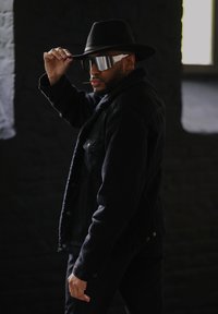 Μαύρο τζάκετ από τζιν με κολάρο από γούνα, μαύρο παντελόνι, καπέλο με φαρδύ γείσο και ασημένια καθρέφτη γυαλιά ηλίου. Στηρίζεται σε σκοτεινό φόντο.