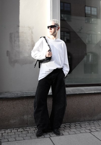 Persona con cabello rubio corto, que lleva gafas de sol negras, camisa blanca de manga larga, pantalones anchos negros y lleva un bolso negro de hombro, está de pie al aire libre.
