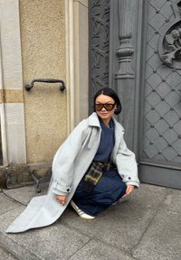 Szary wełniany płaszcz o oversize'owym kroju, granatowy sweter dziany, spódnica dżinsowa i wzorzysty pasek. Duże brązowe okulary przeciwsłoneczne i trampki.