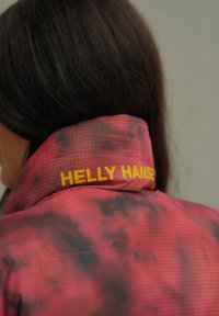 Κόκκινο μπουφάν με σχηματισμό πλέγματος, διαθέτει ψηλό γιακά και κίτρινη επωνυμία "HELly HANSEN" στο πίσω μέρος. Λείο υλικό, στυλ αθλητικών ρούχων.