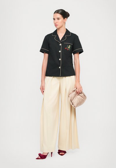 Camisa negra de manga corta con ribetes dorados en contraste y bordado floral, combinada con pantalones de tiro alto en color crema y un bolso beige.