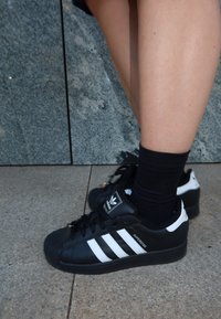 Osoba nosi crne Adidas Superstar tenisice s bijelim prugama i crne čarape, stojeći na kamenoj ploči blizu granitnog zida.