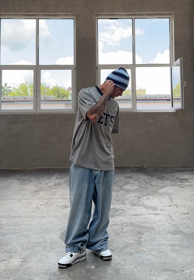 Jersey deportivo gris con el texto "NETS", gorro a rayas azul y blanco, jeans azules holgados y zapatillas blancas con detalles en negro, de pie en interiores.