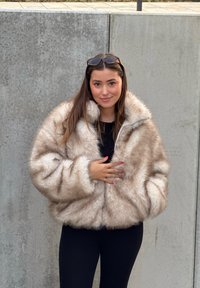 Umetno krzno v svetlo rjavi barvi, z visokim ovratnikom, zapiranjem na zadrgo in volumniziranimi rokavi. Mehka tekstura z gosto, toplo podobo.