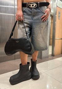 Juodas, kreivai formuotas rankinukas su sidabriniu logotipu, derinamas su šviesiai mėlynomis džinsinėmis šortais ir masyviomis juodomis guminėmis botais su perforacijomis.