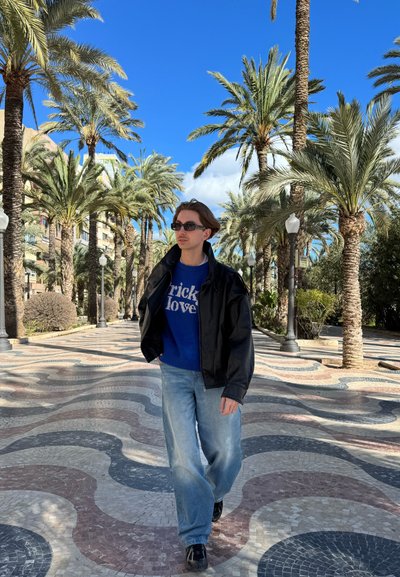 Persona con gafas de sol, chaqueta negra, suéter azul y vaqueros caminando por un paseo con palmeras y baldosas con patrones bajo un cielo azul despejado.