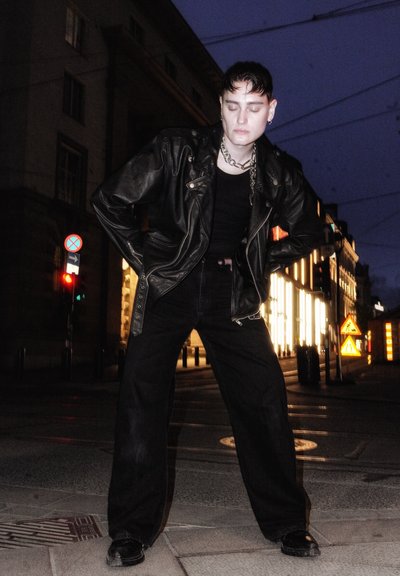 Chaqueta de cuero negra con herrajes plateados, combinada con una camiseta sin mangas negra y pantalones anchos negros. Entorno urbano al atardecer con fondos iluminados.