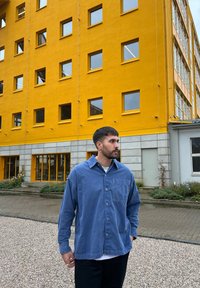 Μακρυμάνικο μπλε πουκάμισο με κουμπιά σε απαλό ύφασμα, με τσέπη στο στήθος, συνδυασμένο με λευκή μπλούζα κι ανδρικό παντελόνι σε σκούρο χρώμα μπροστά από κίτρινο �κτίριο.