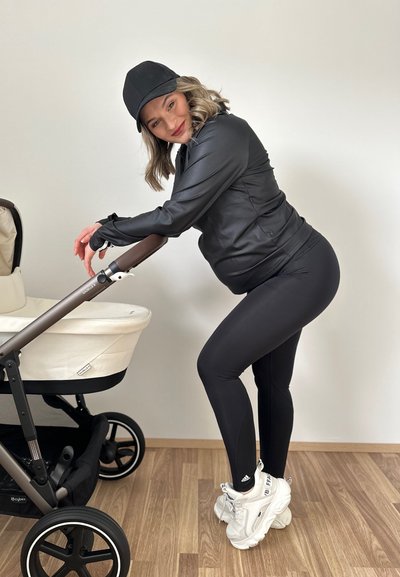 Chaqueta atlética negra y leggings, zapatos blancos, gorra gris; empujando un cochecito de color claro con acentos marrones contra un fondo neutro.