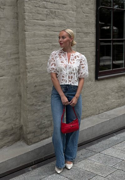 Blusa blanca de encaje con mangas abullonadas, combinada con jeans acampanados de un azul claro. Accesorio: bolso rojo pequeño. El fondo presenta una pared texturizada y una ventana.