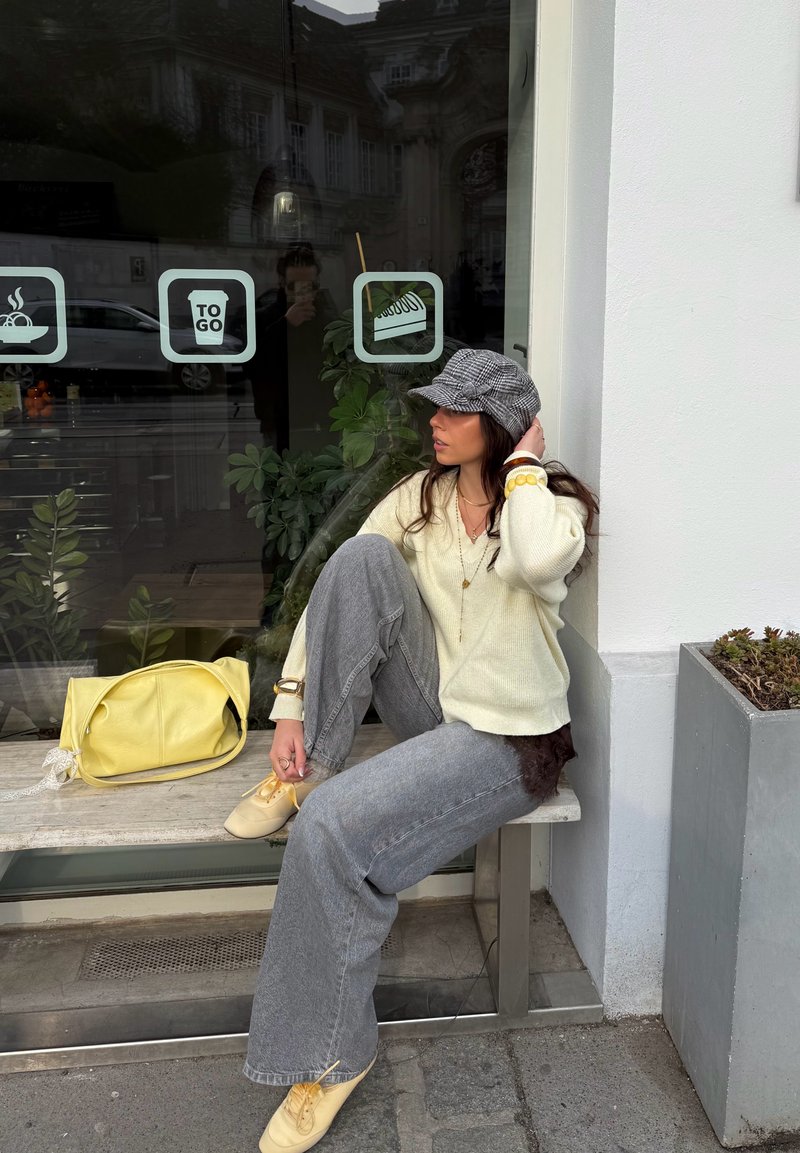 Frau in cremefarbenem Pullover, grauen Jeans und kariertem Hut sitzt auf einer Bank vor einem Café mit einer gelben Tasche auf der Bank neben ihr.