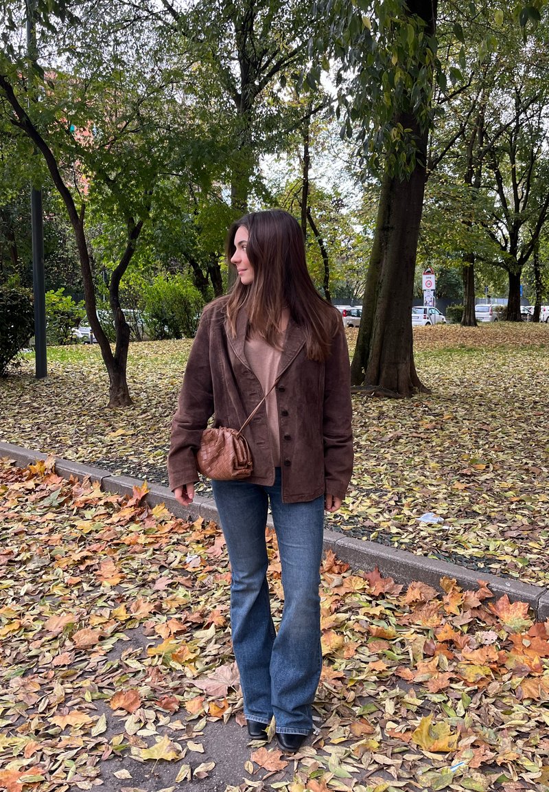 Jachetă din suede maro cu nasturi, top bej, blugi evazați albaștri și o mică geantă din piele matlasată maro, înconjurată de frunze de toamnă.