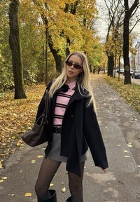 Črn plašč, roza srajca z vzorcem črt, siva krilo, škornji do kolen. Dodatki vključujejo sončna očala in rjavo torbo. Jesensko listje v ozadju.