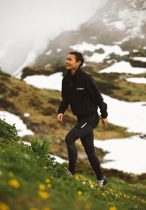 Chaqueta negra Terrex con cuello con cremallera, combinada con leggings negros y grises. La persona está caminando por un terreno cubierto de hierba, rodeada de parches de nieve.