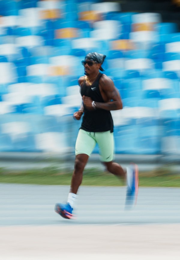 Atleta con camiseta sin mangas negra, pantalones cortos verde claro y zapatos azules corriendo en una pista con asientos vacíos azules y blancos en el estadio de fondo.