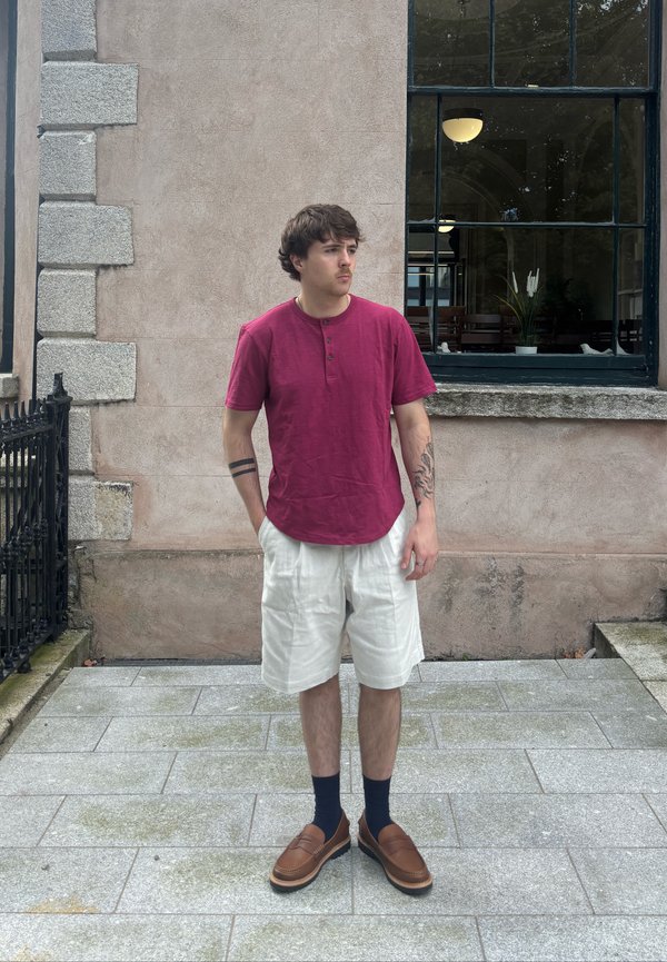 Camiseta henley de mangas curtas bordeaux com três botões, calções creme, mocassins castanhos e meias azuis marinhas contra um fundo de parede de pedra.
