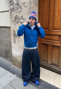 Jeune personne ajustant un sweat à capuche bleu, portant un bonnet coloré, un jean large foncé, une ceinture blanche et des baskets bleues, debout près d'un mur couvert de graffitis et d'une porte en bois.