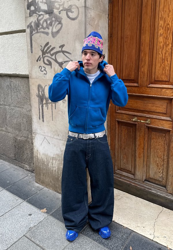 Jeune personne ajustant un sweat à capuche bleu, portant un bonnet coloré, un jean large foncé, une ceinture blanche et des baskets bleues, debout près d’un mur couvert de graffiti et d’une porte en bois.