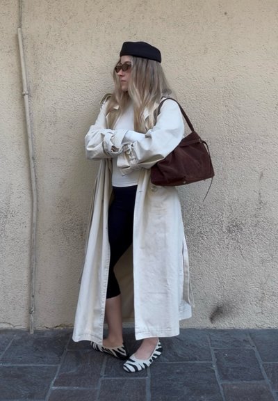 Mujer que lleva un abrigo beige, boina negra, gafas de sol, leggings negros, zapatos planos con estampado de cebra y lleva un bolso marrón al hombro, apoyada contra una pared.