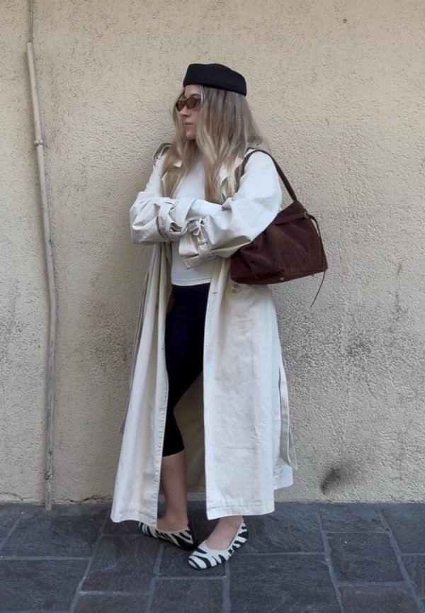 Kvinne iført en beige trenchcoat, svart beret, solbriller, svarte leggings, flate sko med zebramønster, og bærer en brun skulderveske, stående mot en vegg.