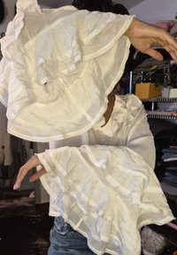 Fehér, könnyű blúz fodros ujjakkal, vízszintes csíkokkal és enyhén áttetsző textúrával. Látható kéz- és karmozdulatok.