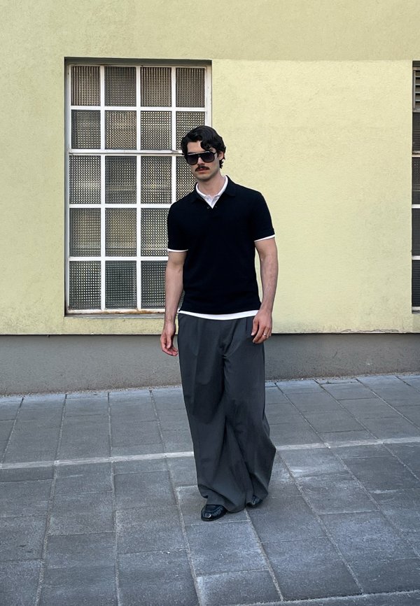 Άνδρας με μουστάκι που φοράει μαύρα γυαλιά ηλίου, μαύρο πόλο μπλουζάκι και φαρδιά γκρι παντελόνια περπατά σε γκρι πλακόστρωτο πεζοδρόμιο δίπλα σε κίτρινο τοίχο.