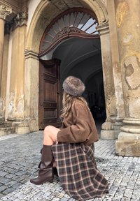 Pruun seemisjakk, tumedates toonides ruuduline seelik, põlveni ulatuvad pruuni värvi saapad ja karvamüts; istub munakivitänaval puust ukse lähedal.