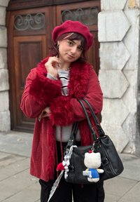 Veste en velours côtelé rouge avec bordure en fourrure, pull rayé gris, sac à main noir avec porte-clés en peluche, et béret bordeaux foncé. Fond en pierre.