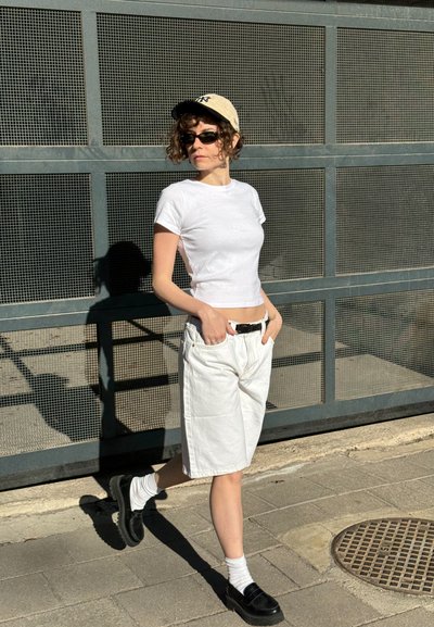 Camiseta ajustada de manga corta blanca, pantalones cortos de denim blancos, zapatos negros con cordones, calcetines cortos blancos, gorra beige, gafas de sol. Fondo texturizado.