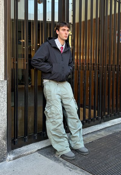 Chaqueta negra con capucha, camisa de cuadros y corbata roja. Pantalones cargo verdes claros y holgados con bolsillos. Botas grises. De pie contra barrotes de metal.