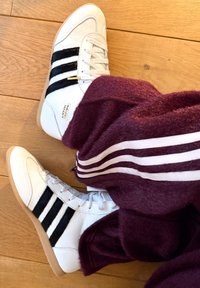 Baskets blanches Adidas Japan avec des bandes noires, portées avec un pantalon bordeaux à bandes blanches latérales, reposant sur un sol en bois.