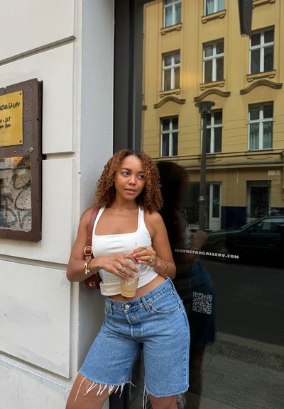 Top sin mangas blanco de escote cuadrado, pantalones cortos de mezclilla de cintura alta con dobladillo deshilachado, sosteniendo un café helado. Al fondo se ve una ventana de galería y un cartel amarillo.