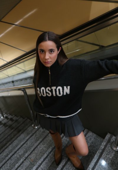 Suéter negro con "BOSTON" en blanco, cuello alto, cierre plateado, combinado con una falda gris con pliegues y botas marrones hasta la rodilla, en unas escaleras.