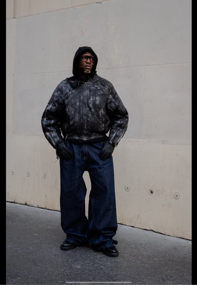Chaqueta de cuero negra texturizada con cremallera, pantalones vaqueros de mezclilla oscura de corte holgado, guantes negros y zapatos. De pie contra un fondo de pared neutro.