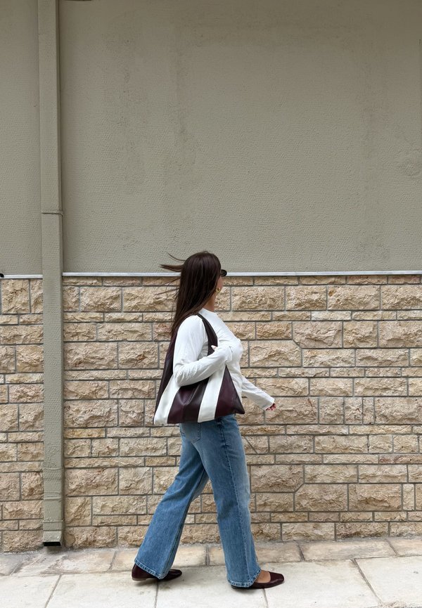 Mujer con cabello largo y oscuro, que lleva gafas de sol, camiseta blanca, vaqueros azules y zapatos marrones, caminando junto a una pared de piedra beige y llevando un bolso de hombro bicolor.