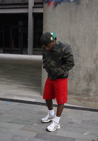 Camo-jas, rode shorts en witte sportschoenen. De jas heeft een rits en zakken. De pet heeft een cirkelvormig logo op de voorkant.