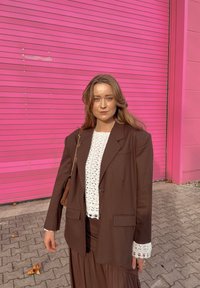 Blazer marron oversized sur un haut en maille blanc, associé à une jupe marron. Accessoires : un petit sac marron et des bijoux en or. Fond rose.