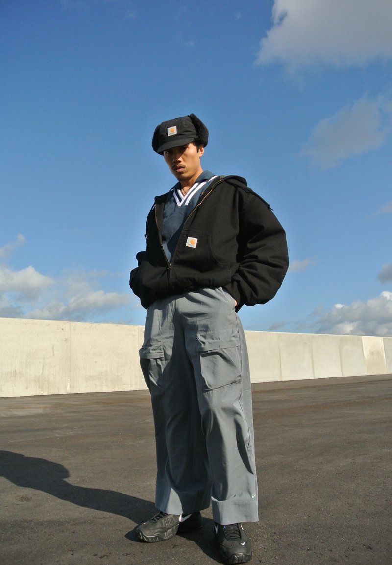 Μαύρη φούτερ με κουκούλα και μεγάλα τσέπες, ανοιχτό γκρι cargo παντελόνι και μαύρα αθλητικά παπούτσια, με φόντο έναν μπλε ουρανό με σύννεφα.